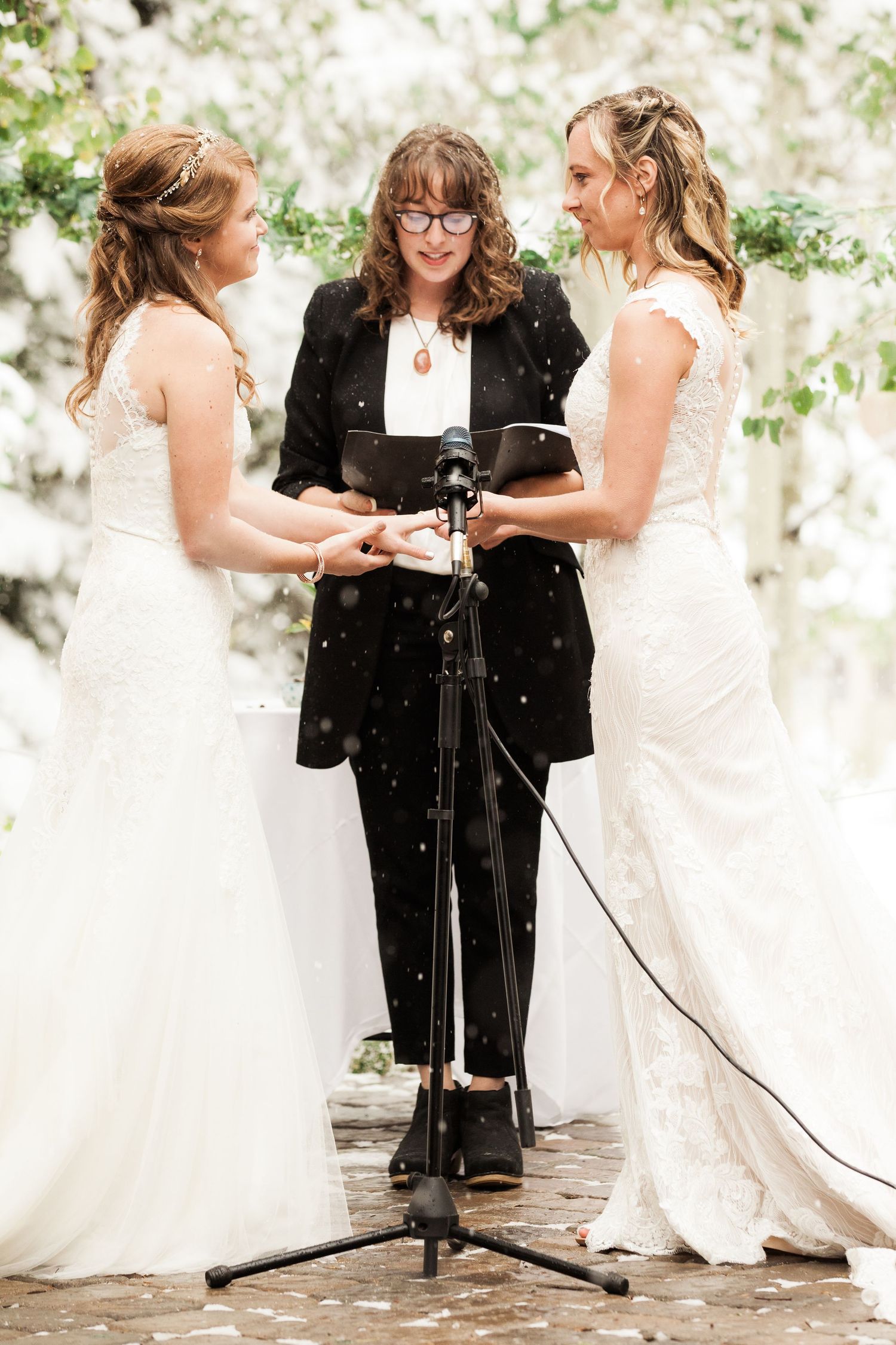 A wedding ceremony takes place outdoors as two brides in white dresses exchange vows with an officiant in black.