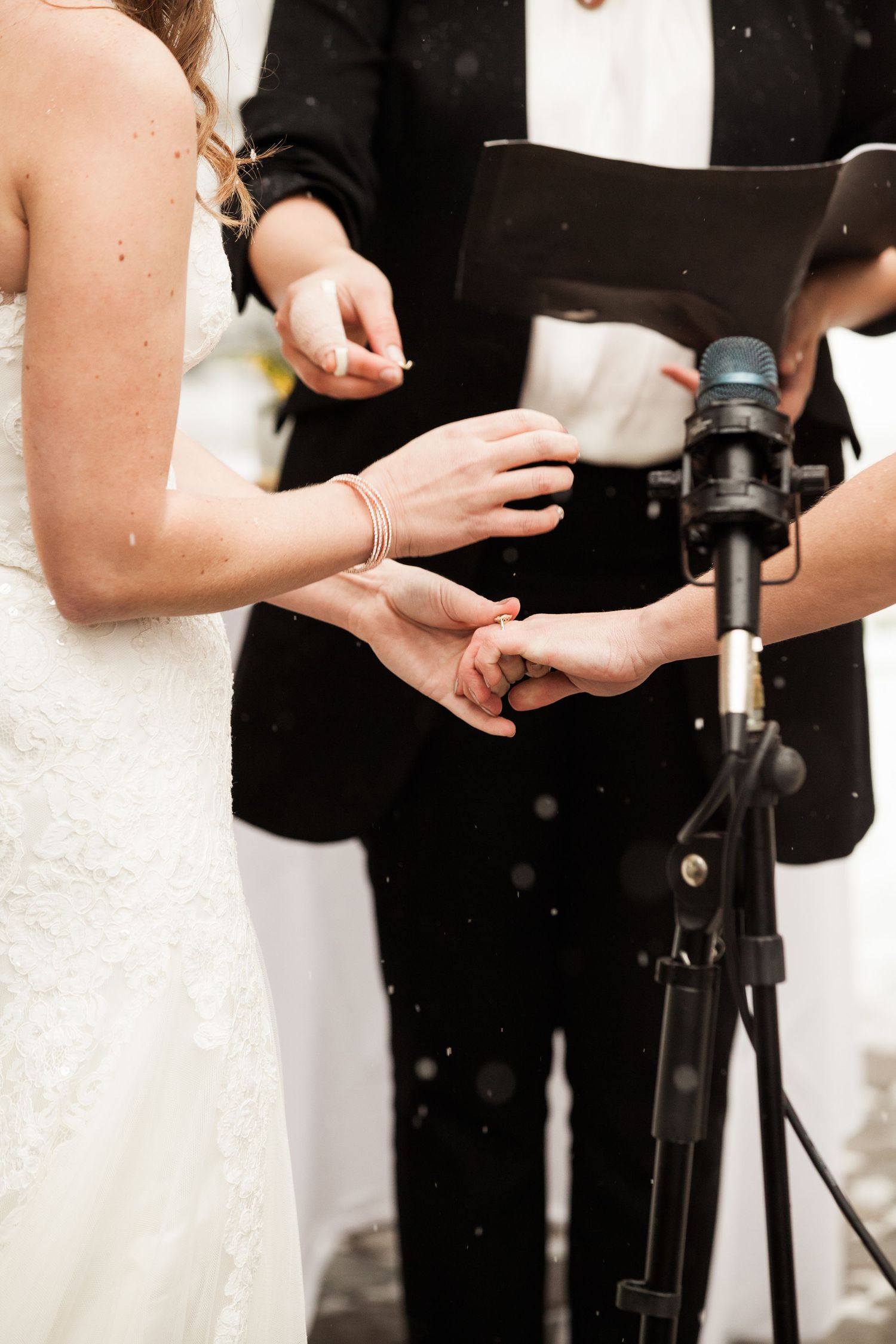 A close-up of hands and microphone during a wedding ceremony with bride in white dress and officiant in black.