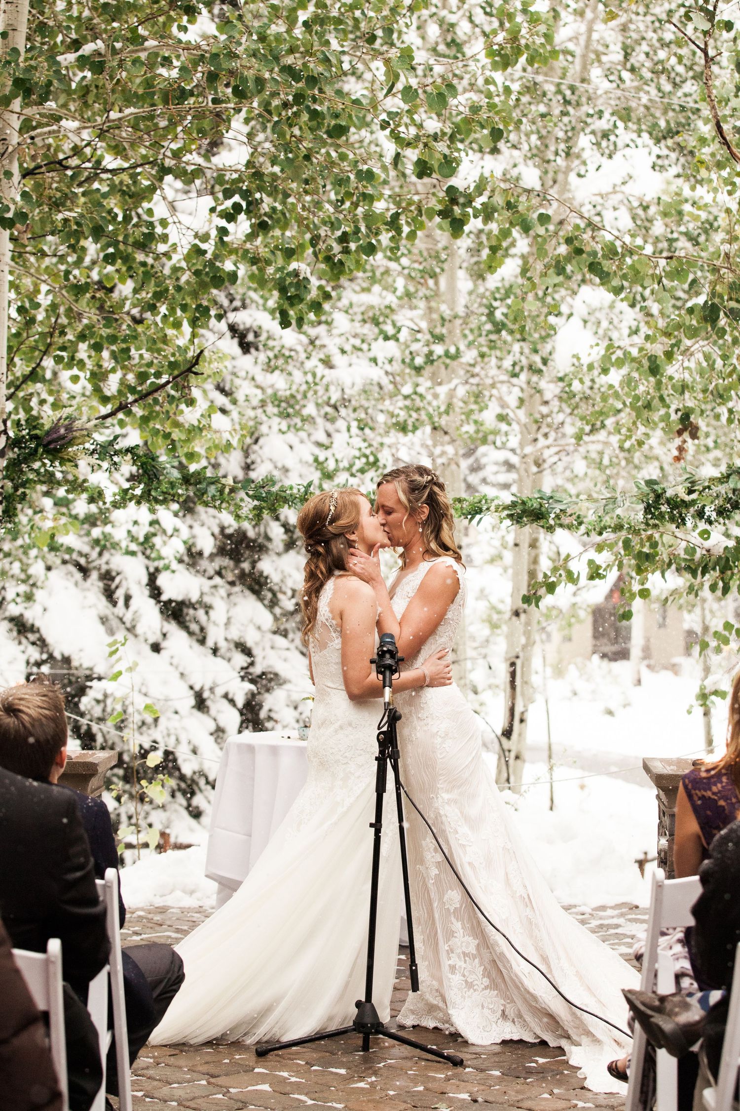 Two brides share a romantic moment during their outdoor wedding ceremony under leafy trees.