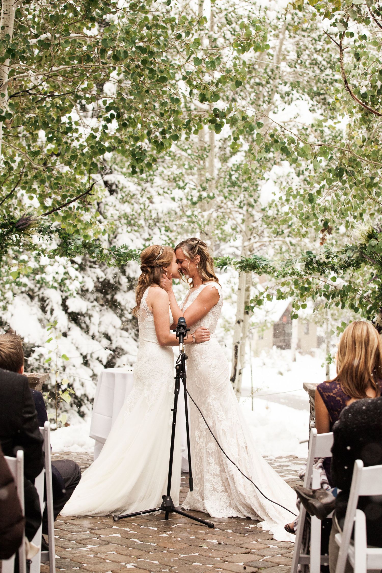 A beautiful outdoor wedding ceremony under a canopy of trees with guests seated on white chairs on a stone path.