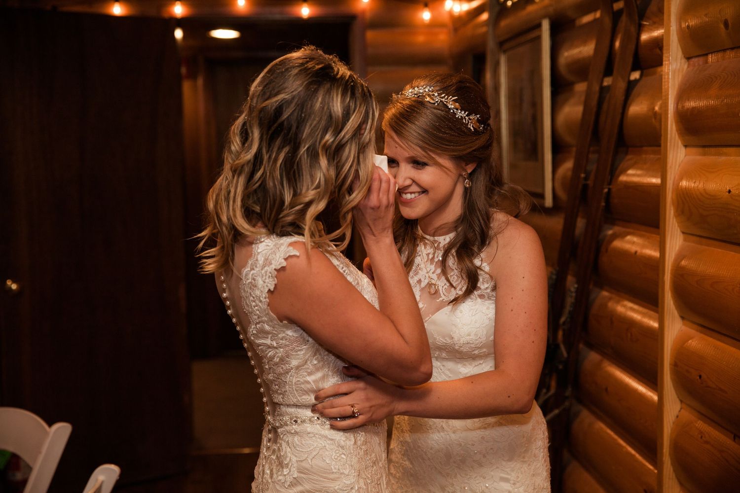 Two brides share an intimate emotional moment in a rustic log cabin venue during their wedding celebration.