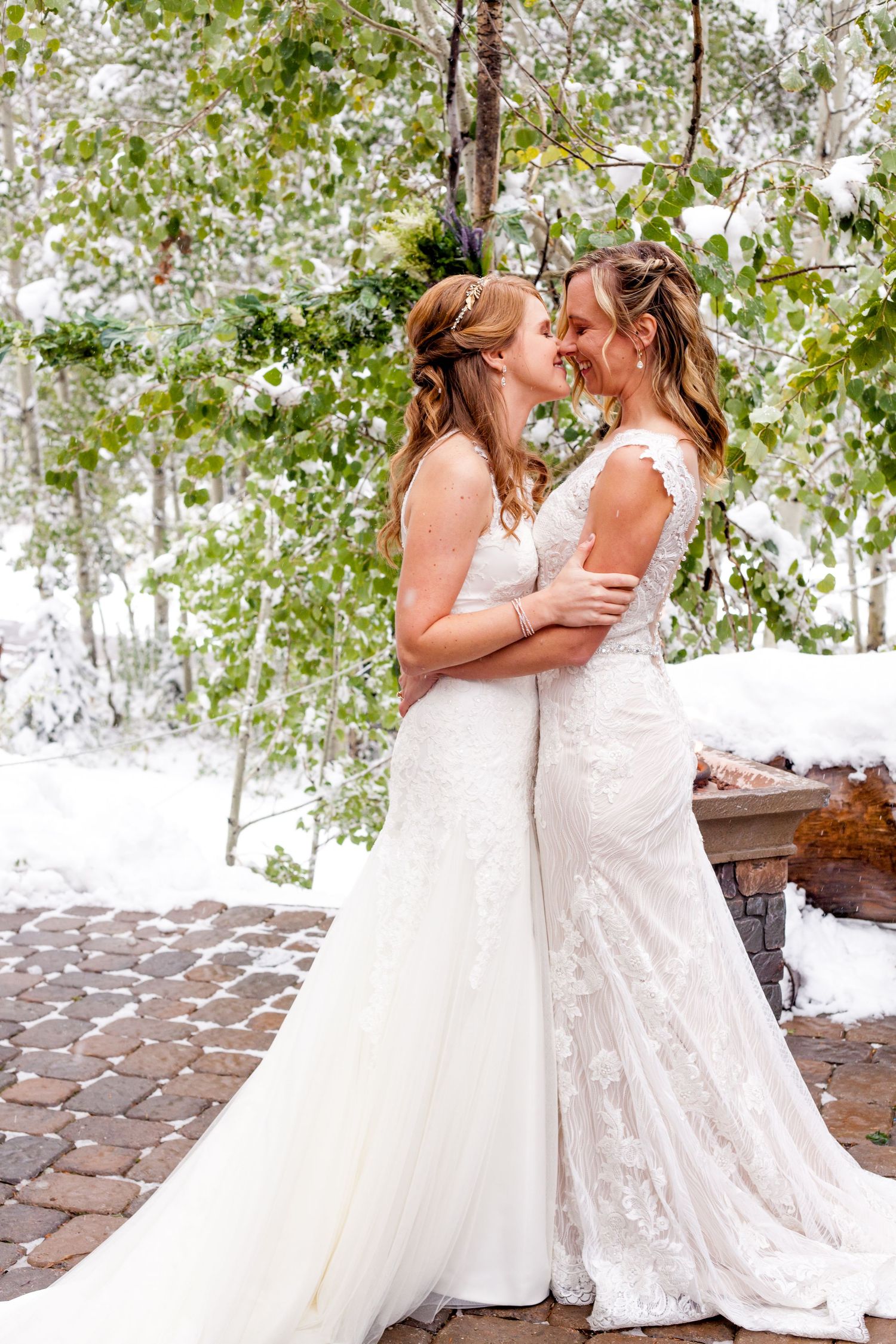 Two brides in elegant white wedding dresses share a joyful moment together in a snowy outdoor setting with trees.