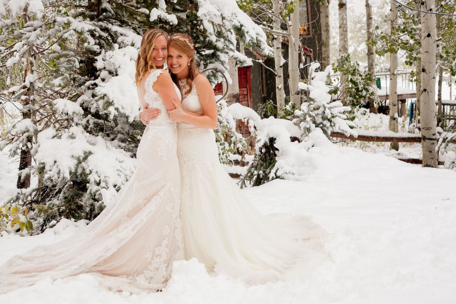 Two brides embrace romantically in winter wedding dresses against a snowy forest backdrop.