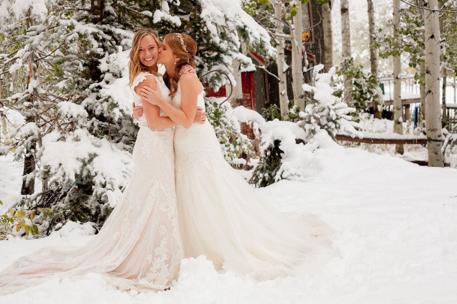 Couple embraces in their wedding dresses against a snowy winter backdrop.