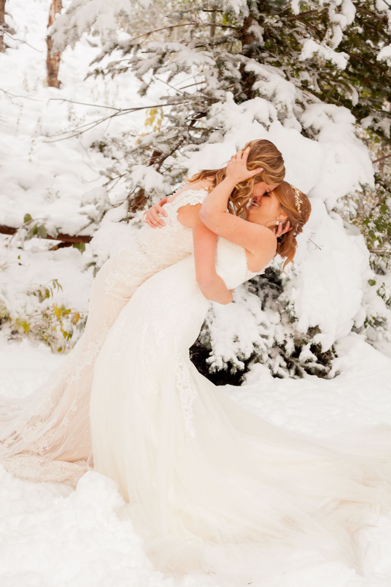 A romantic winter wedding scene with a couple embracing in their white gown and suit among snow-covered evergreen trees.