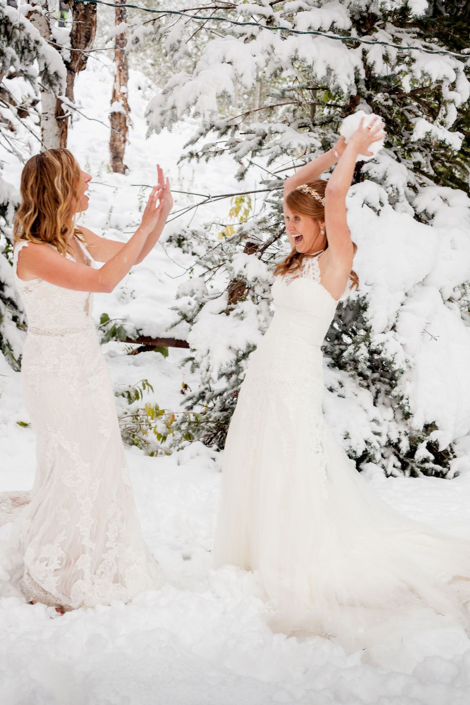 Two women in white wedding dresses playfully throwing snow at each other in a winter wonderland forest setting.