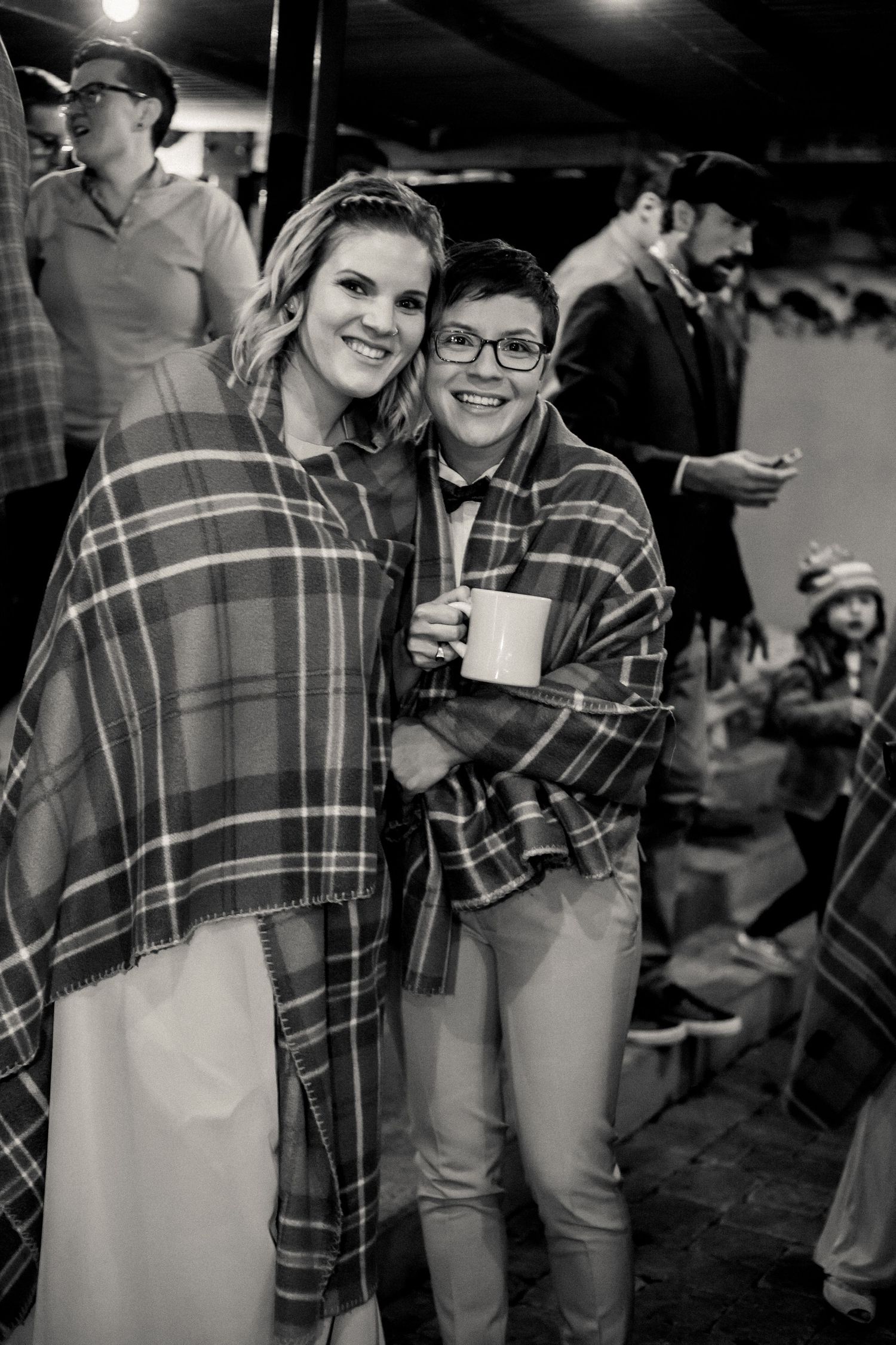 Two people wrapped in plaid blankets smile while holding drinks at an outdoor gathering in black and white.