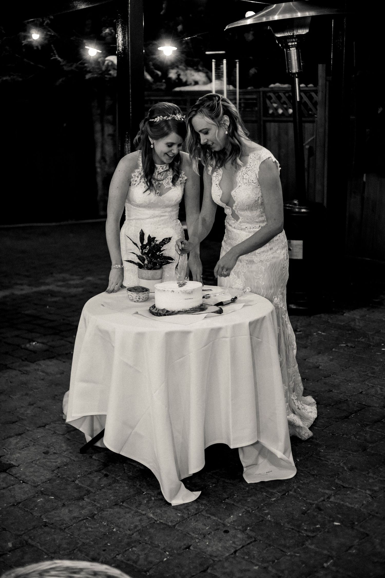 A black and white photo of two brides in white dresses sharing a moment at their wedding cake table.
