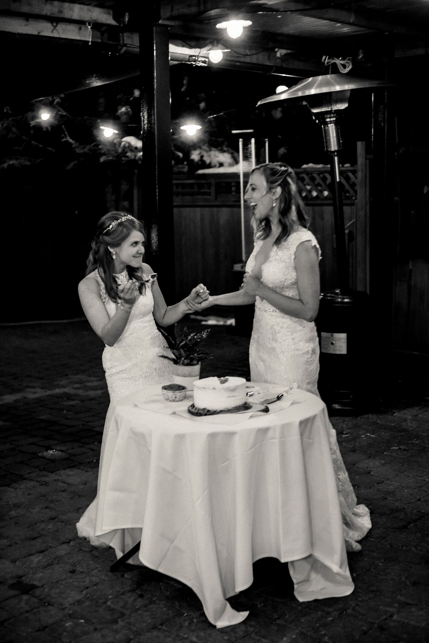 Two people in white dresses share a sweet moment while cutting cake together at a rustic reception venue.