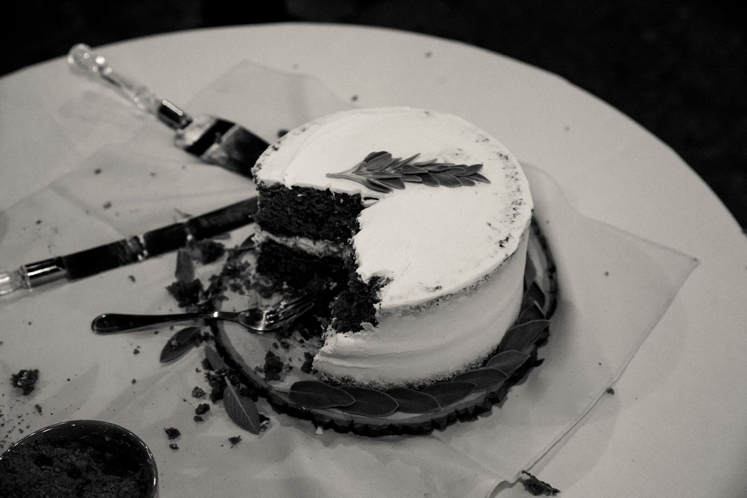A black and white photograph of a sliced chocolate cake with creamy white frosting and dark crumbs on a plate.