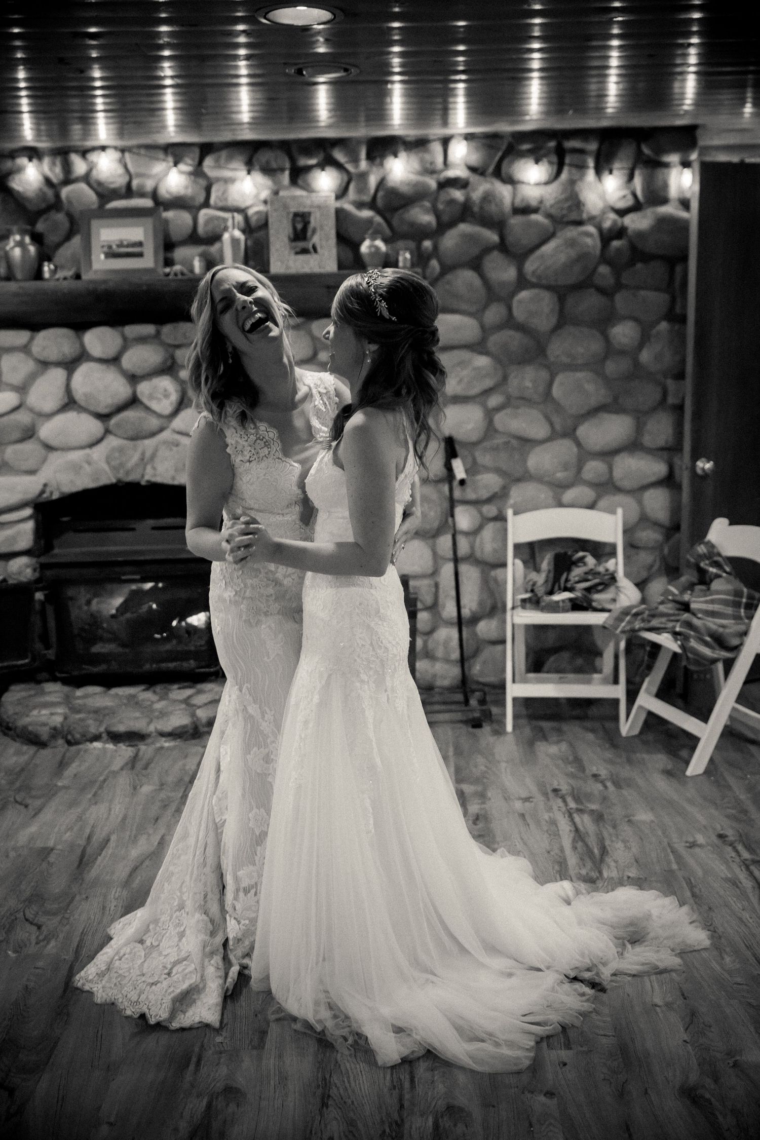 A romantic wedding moment captures two brides in white lace gowns dancing together on a hardwood floor with stone walls behind them.