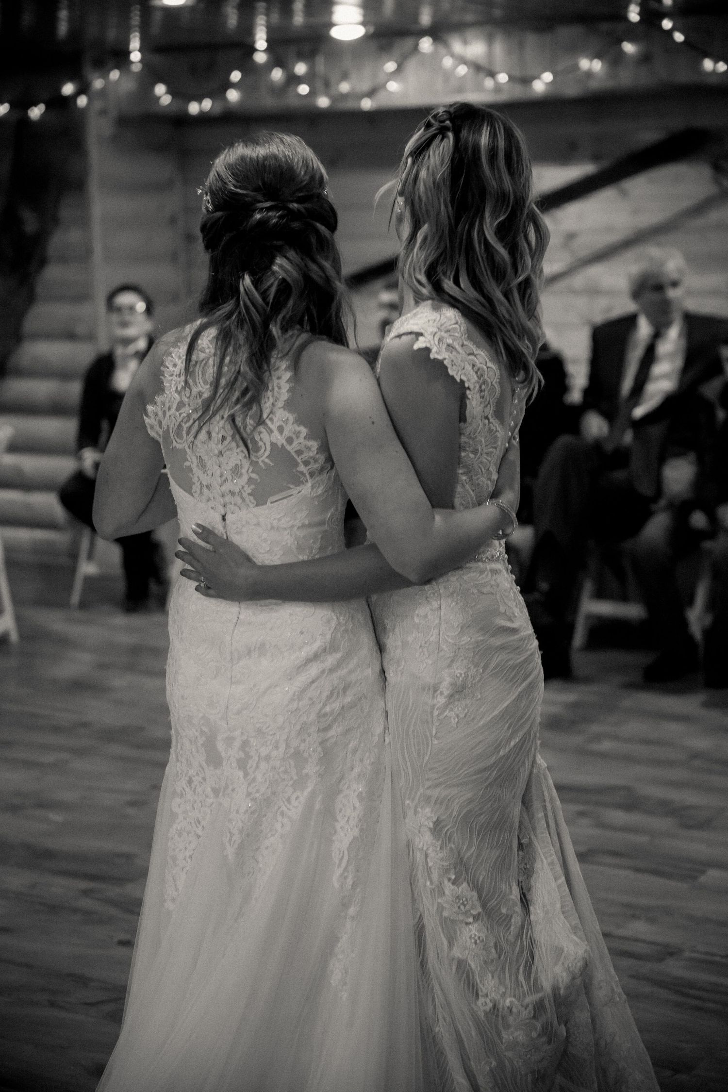 Two brides in elegant lace wedding dresses embrace while dancing at their wedding reception in black and white.