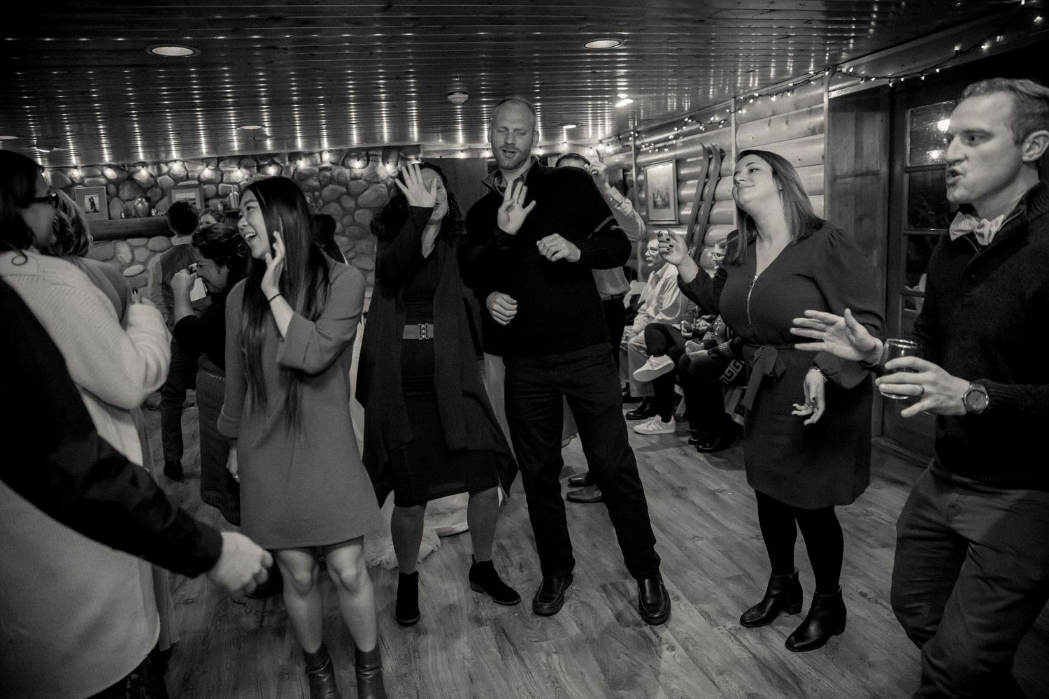 A group of people dancing and socializing at a party in a dimly lit room with wooden floors and ceiling lights.