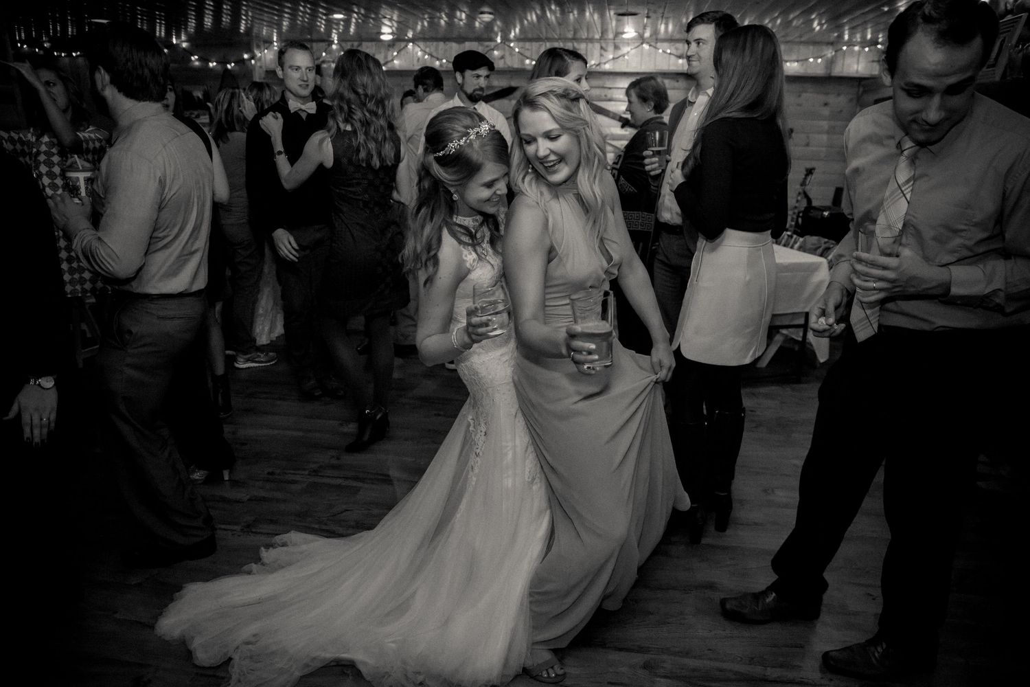 Two women dance together on a crowded dance floor during a wedding reception, one in a bridal gown and one in a formal dress.