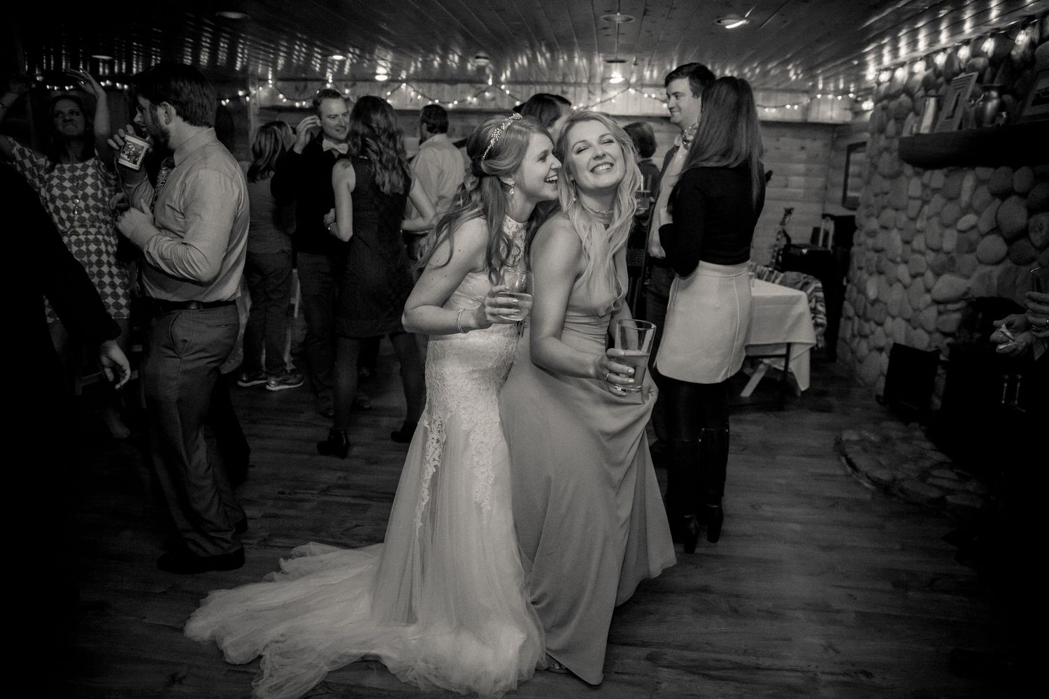 Black and white photo of wedding guests dancing and celebrating together on a wooden dance floor at a reception.