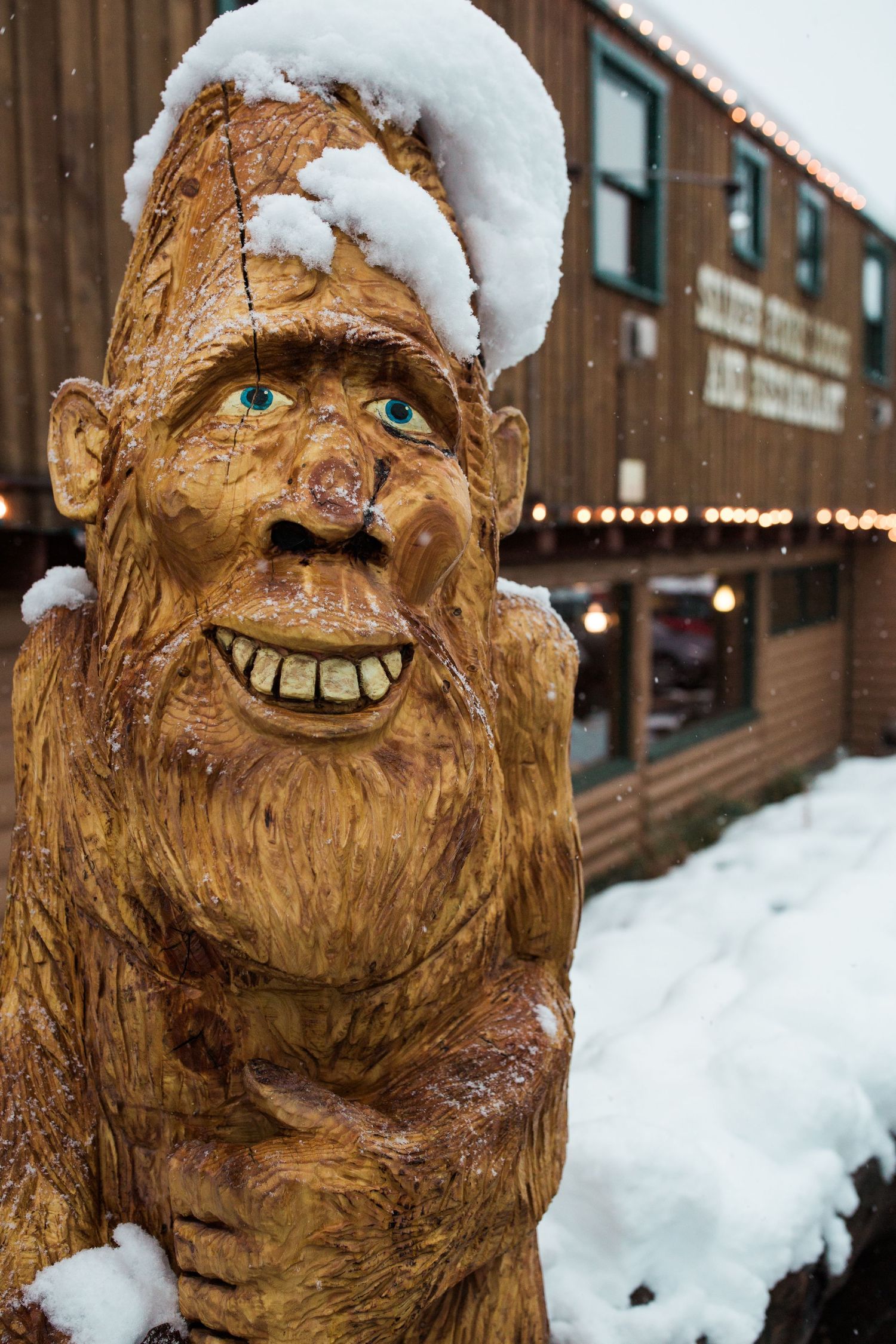 A carved wooden troll statue with snow on its head grins outside a rustic building in winter.
