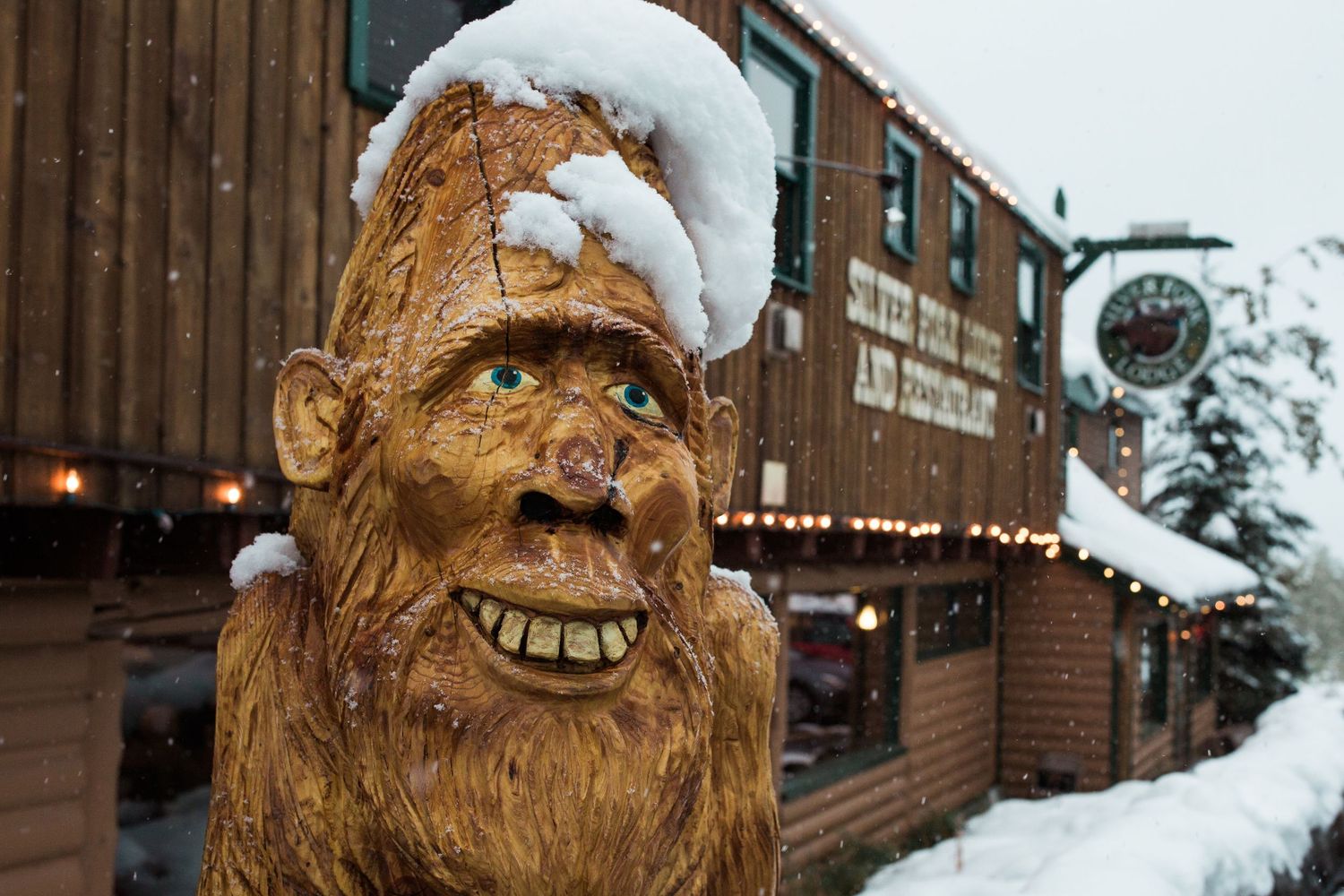 A snow-covered wooden sculpture of a bearded face with a big grin outside a rustic mountain lodge.