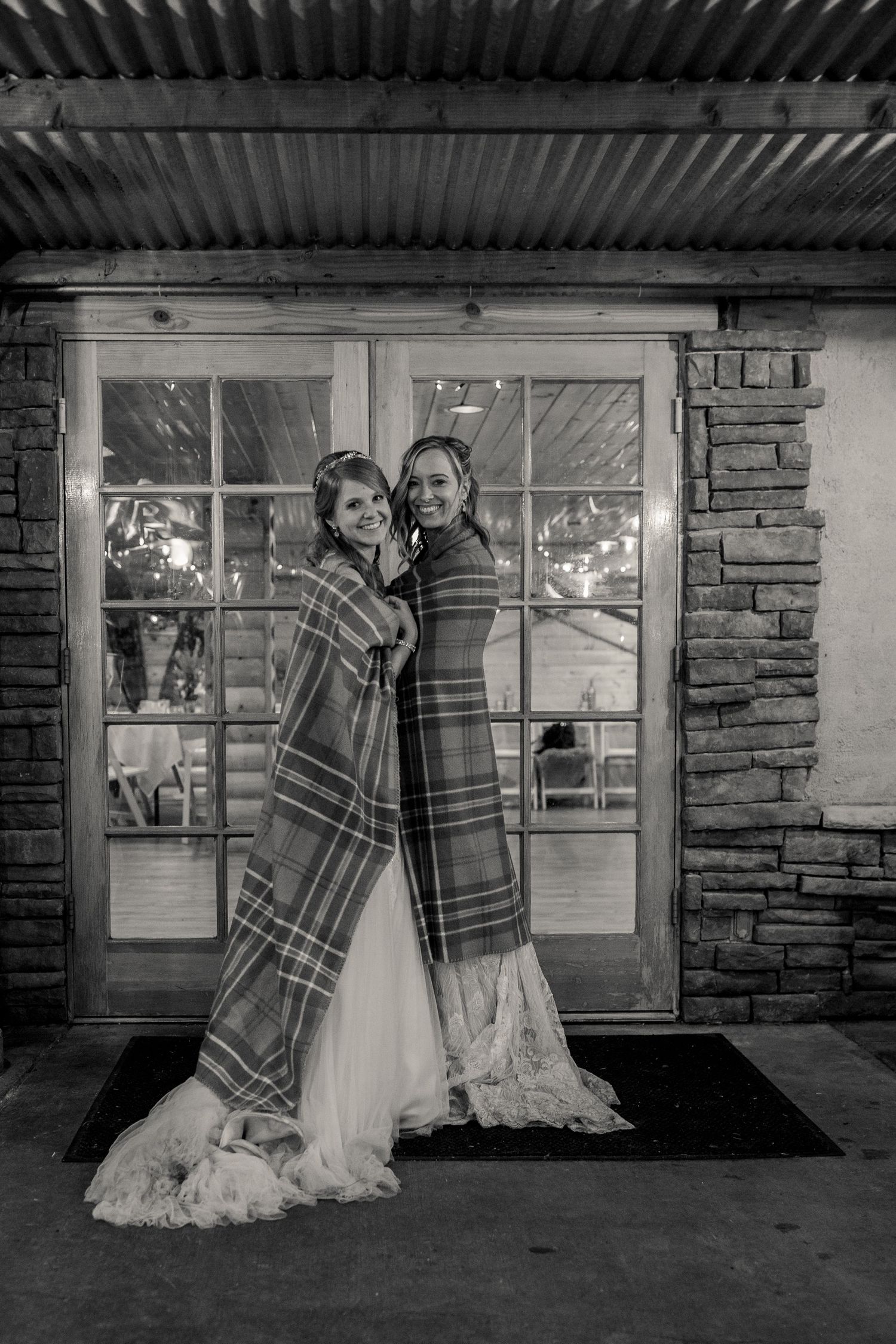 Two people in wedding gowns share a plaid blanket while standing together on a porch in black and white.