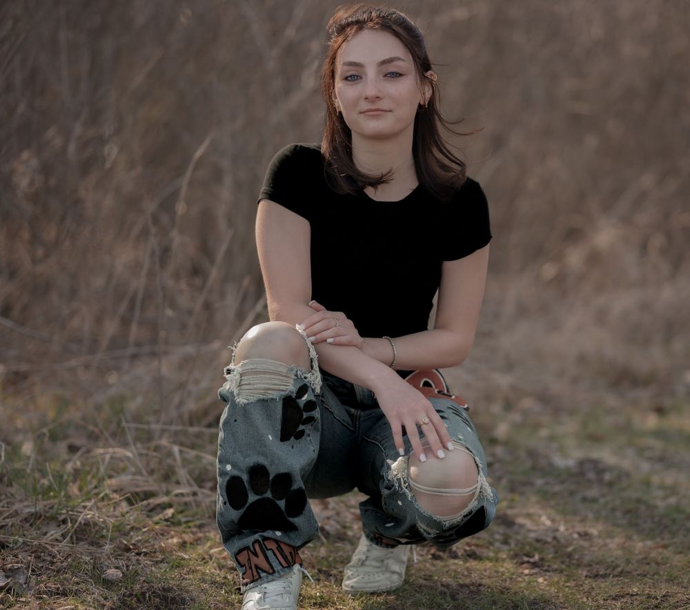 Person in a black t-shirt and ripped jeans crouches outdoors in a natural setting during golden hour.