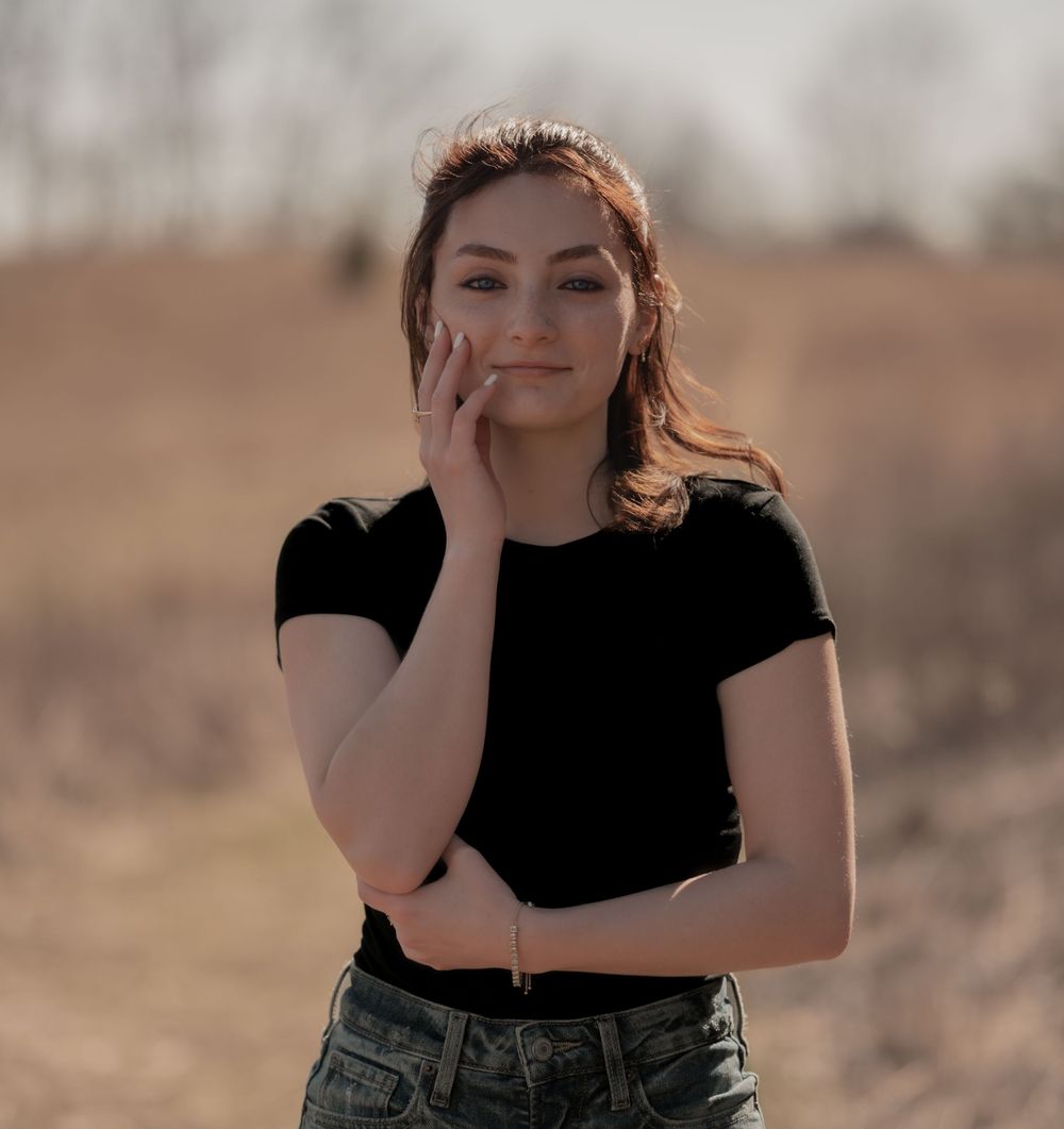 Portrait photo outdoors in desert landscape wearing black t-shirt and denim with hand resting on cheek.