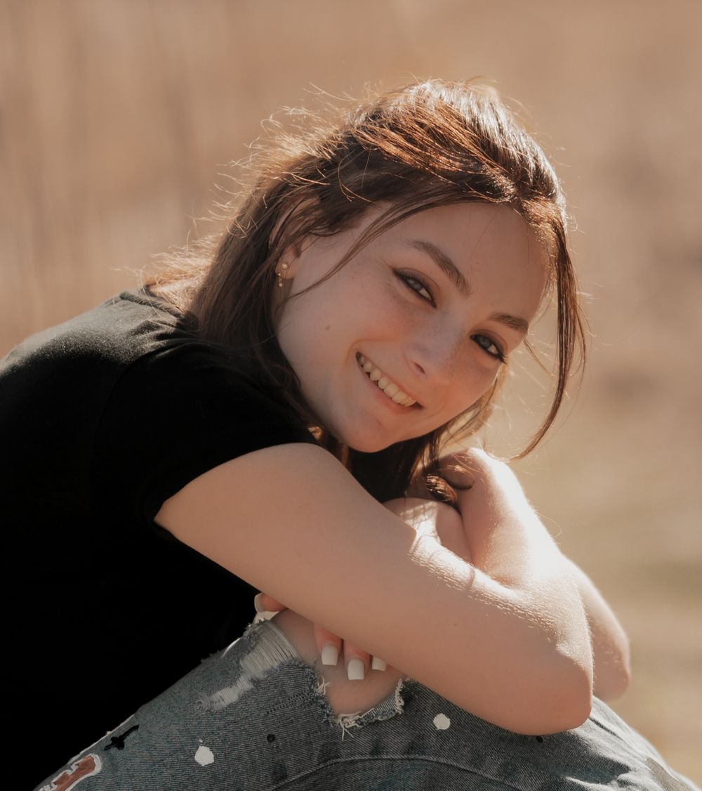 A person in a black sweater smiles warmly while resting their head on their arm in soft sunlit outdoor setting.