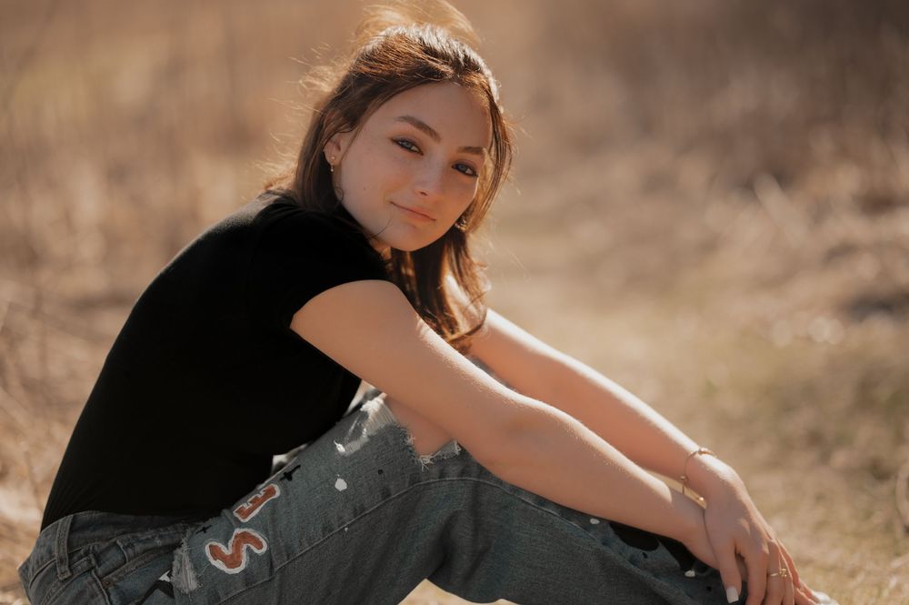A person in black top and jeans sits casually on the ground in warm natural lighting.