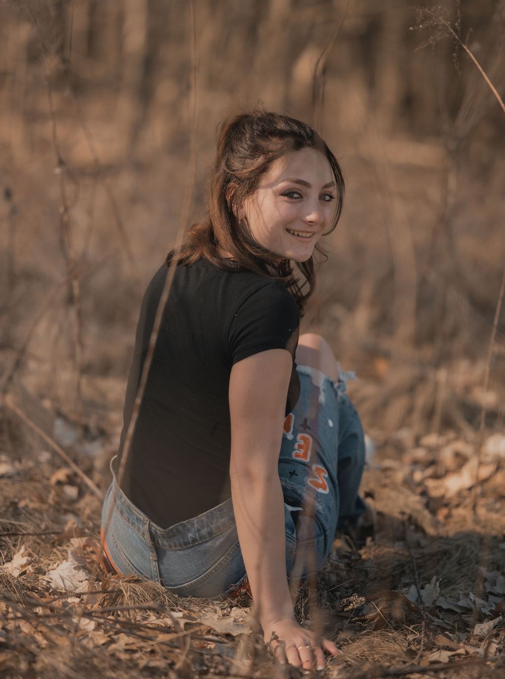 Person in black shirt and jeans sitting on ground while looking over shoulder in warm lighting.
