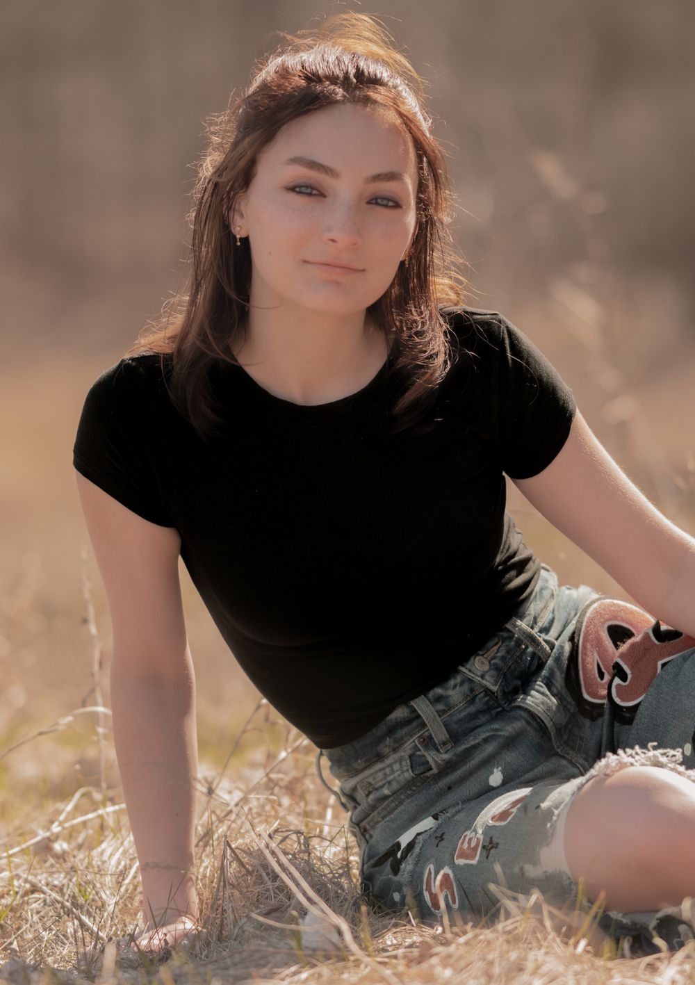 A person in a black t-shirt and denim shorts sits in a sunny field with dried grass during golden hour.