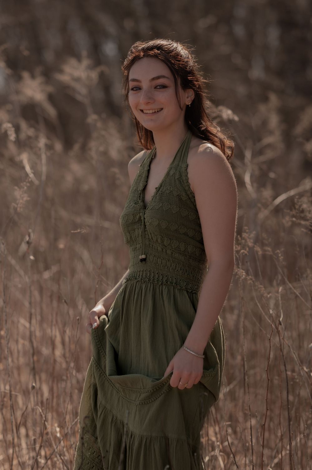 A person in an olive green halter dress poses gracefully in a sun-lit field of tall grass during golden hour.