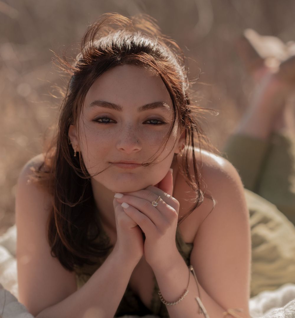A young woman in a natural sunlit setting rests her head on clasped hands while smiling softly at the camera.