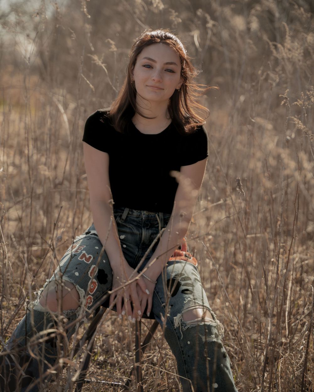 Portrait of a person in a black t-shirt and distressed jeans sitting in a field of tall grass at sunset.