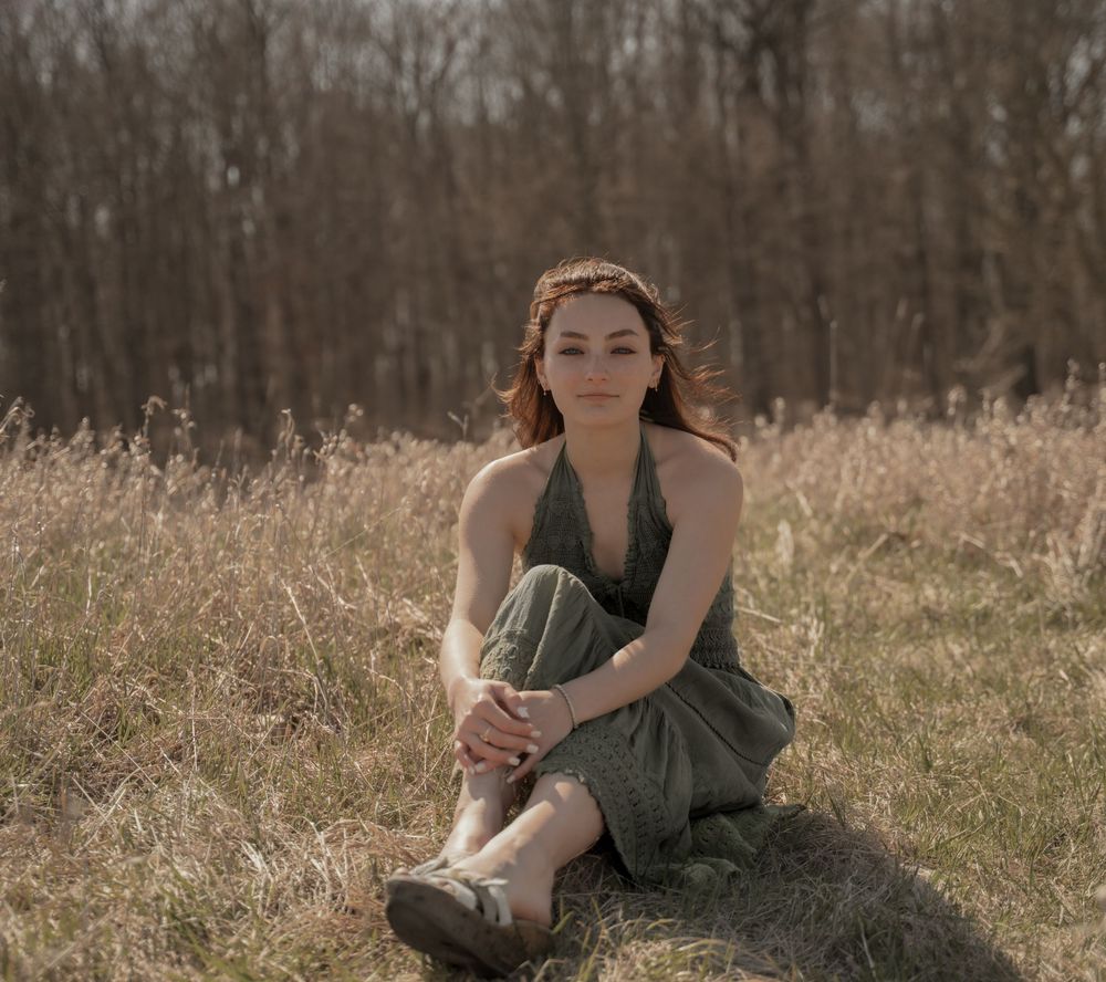 A person in a green dress sits thoughtfully in a sunlit meadow with autumn trees in the background.