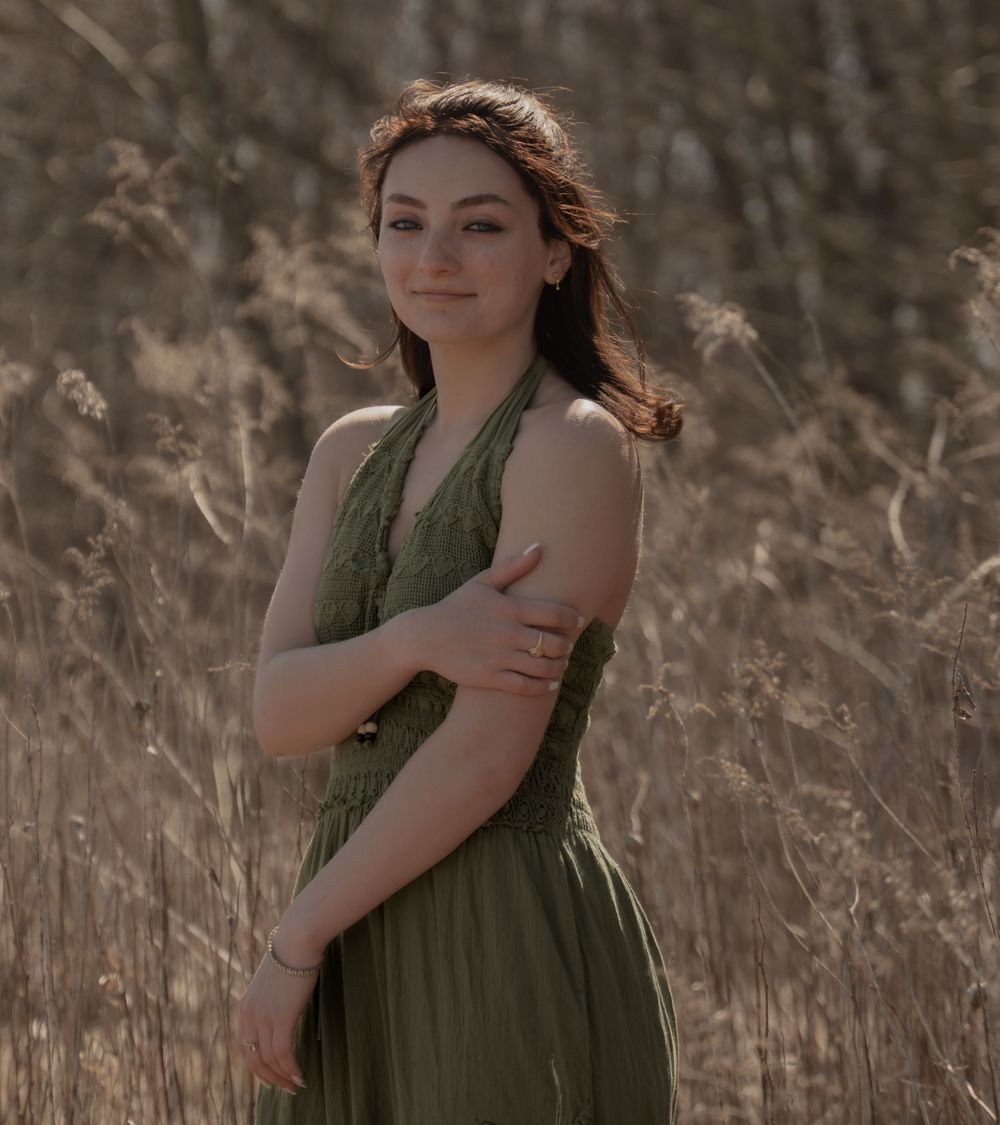 A person in a flowing olive green dress poses thoughtfully in a sunlit field of dried grass and reeds.