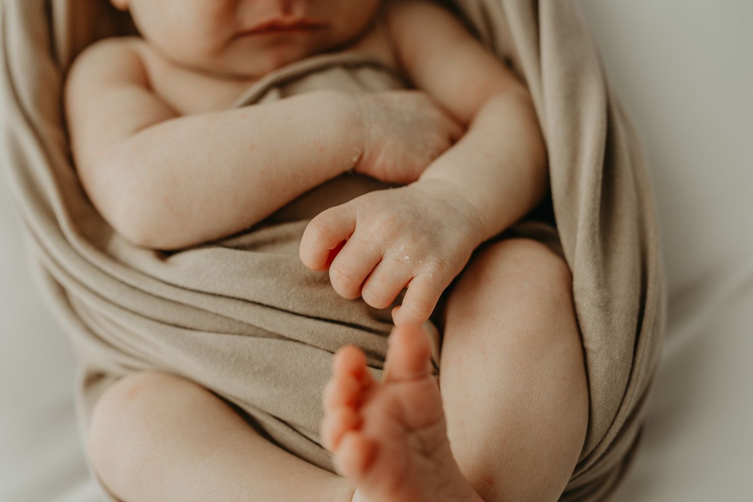 Close-up of newborn baby's feet and hands wrapped in soft beige fabric against neutral background.