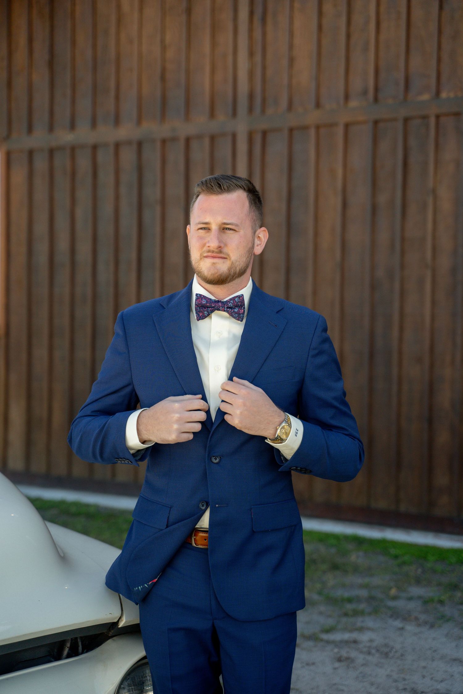 Person in navy blue suit and bow tie standing next to vintage car.