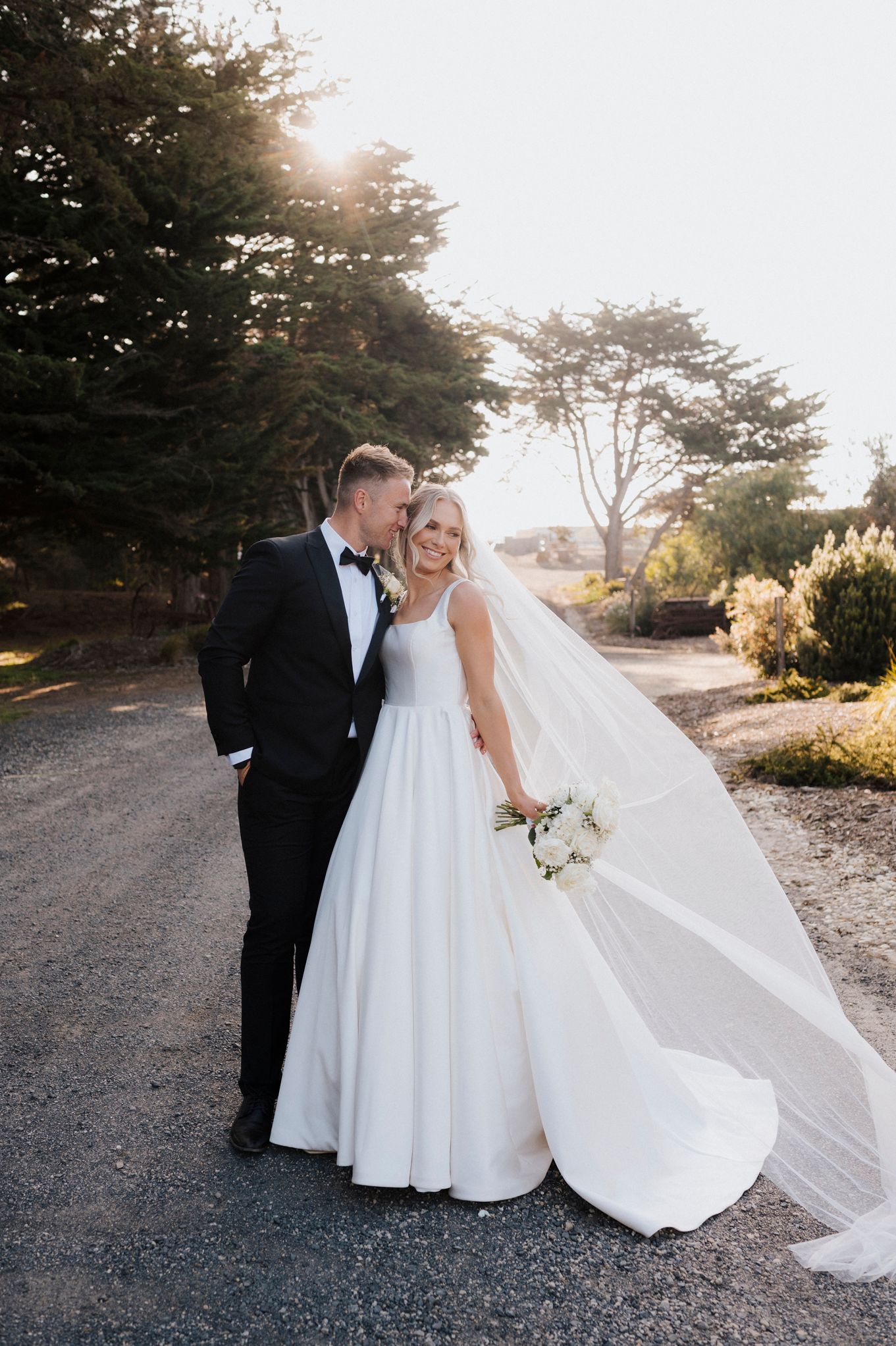 Wedding couple poses together on a path at sunset with bride's flowing veil creating elegant silhouettes.