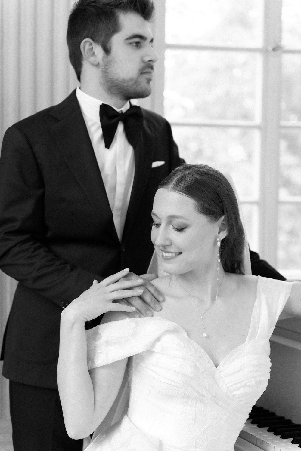Black and white portrait of a elegant couple in formal wedding attire posing near a piano in an intimate setting.