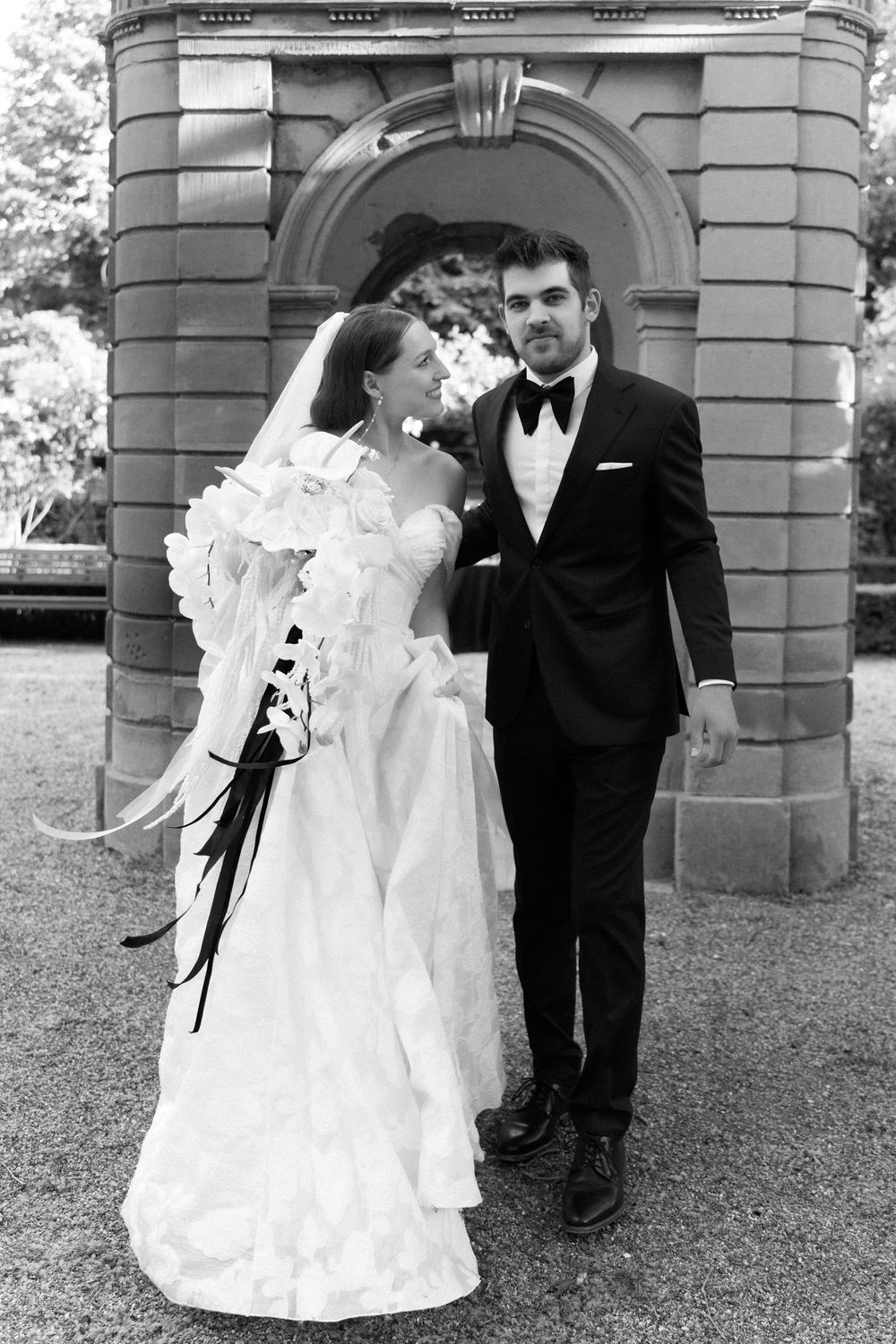 A black and white wedding photograph showing a bride in a vintage white dress and groom in tuxedo standing by stone archway.