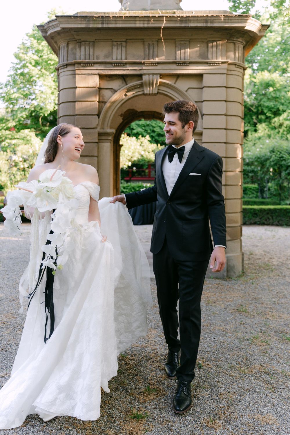 A bride in a white off-shoulder gown and groom in black tuxedo share a joyful moment by a stone archway in a garden setting.