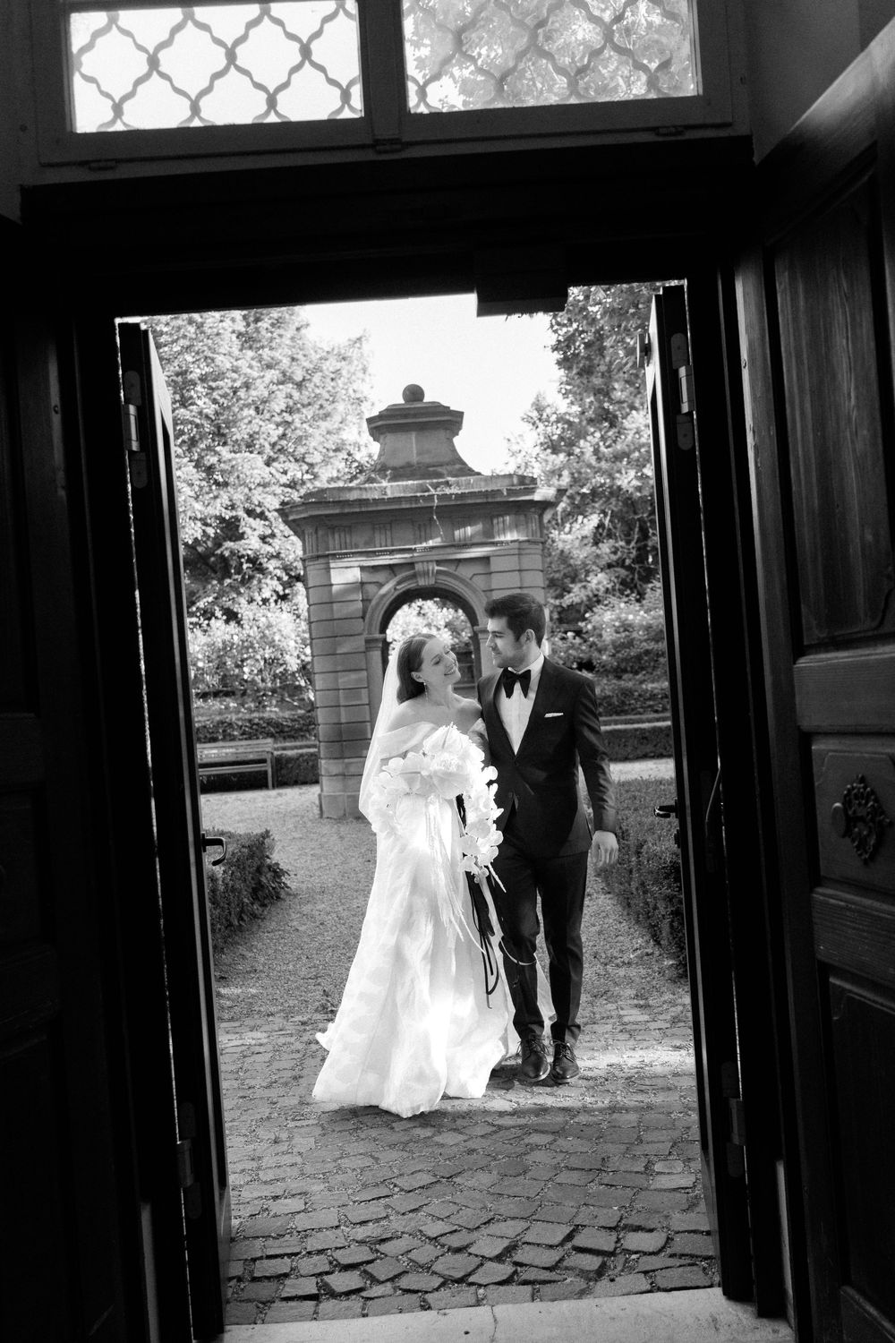 Black and white wedding photo framed through an ornate doorway showing a couple on cobblestone path by stone pillar.