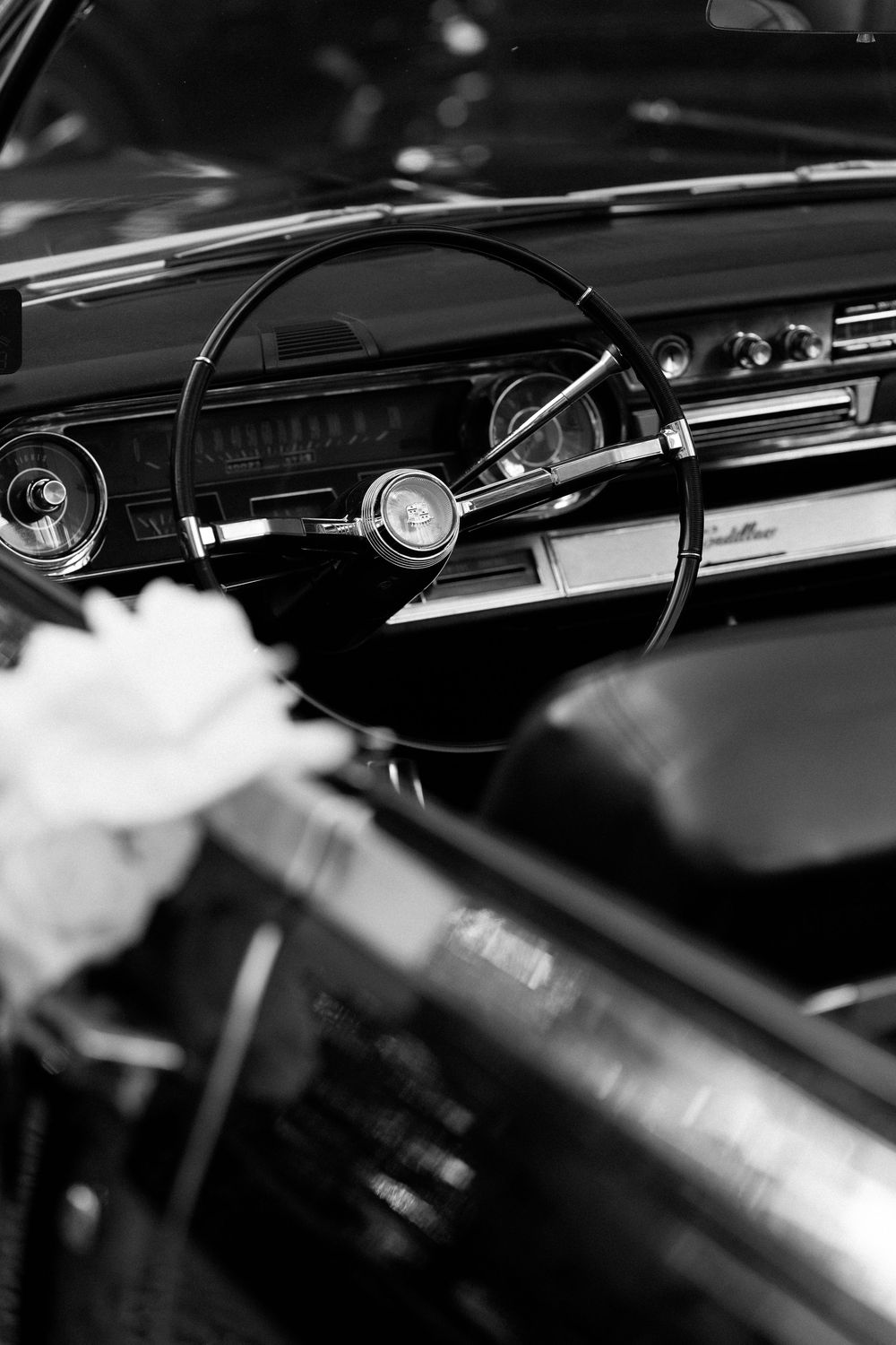 Black and white close-up of a vintage car steering wheel and dashboard with chrome details and circular gauges.