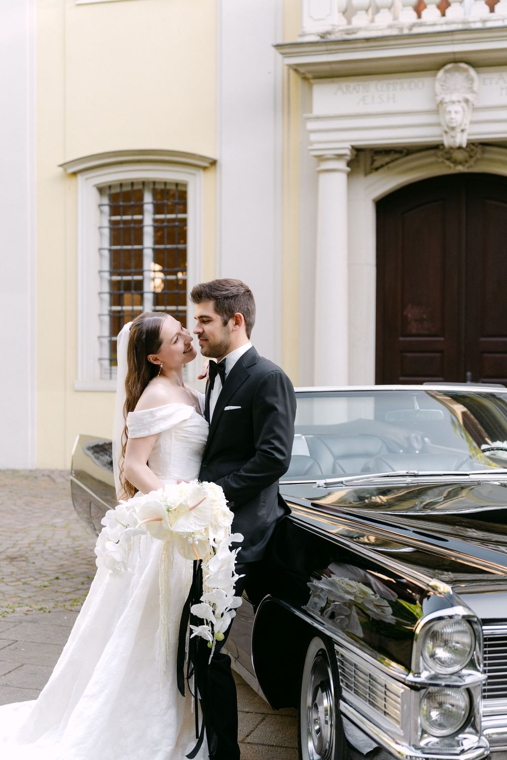 A romantic couple embraces by a vintage black Cadillac outside an elegant historic building on their wedding day.