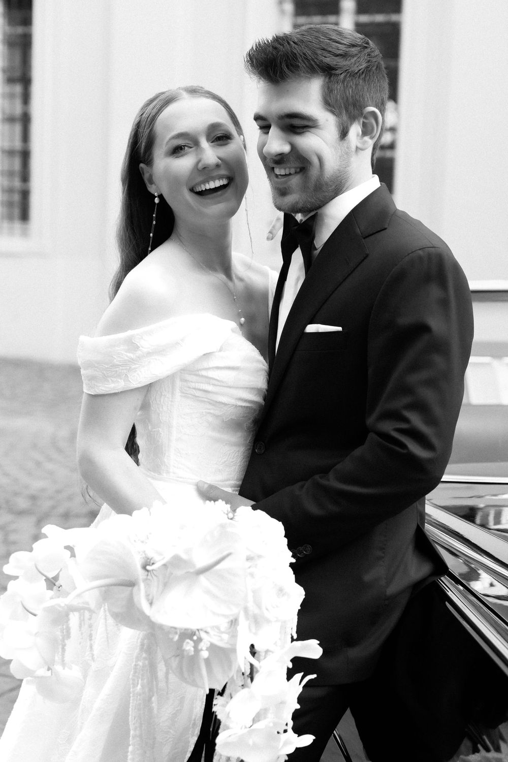 A black and white wedding portrait showing a joyful couple, with the bride in an off-shoulder gown holding a white bouquet.
