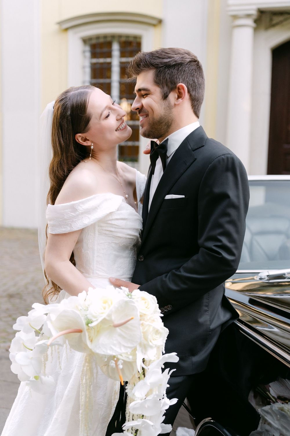 A romantic newlywed couple share a tender moment next to a vintage car while holding a white floral bouquet.