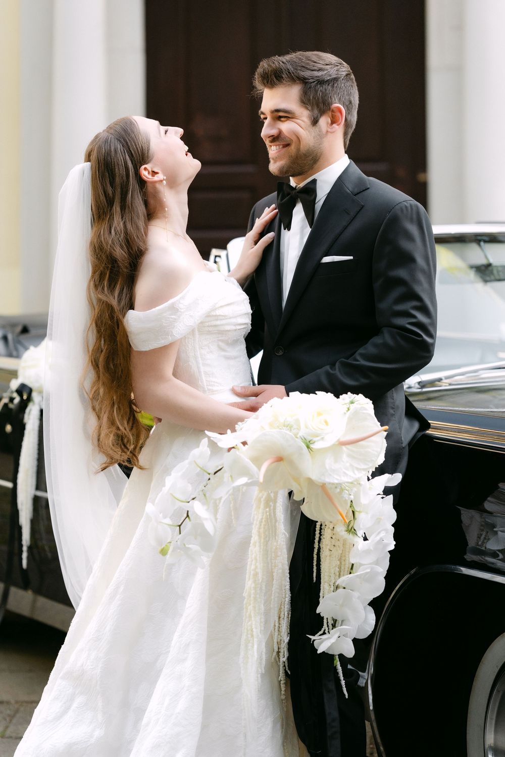 Newlywed couple share a joyful moment beside vintage car, bride holding cascading white orchid bouquet.