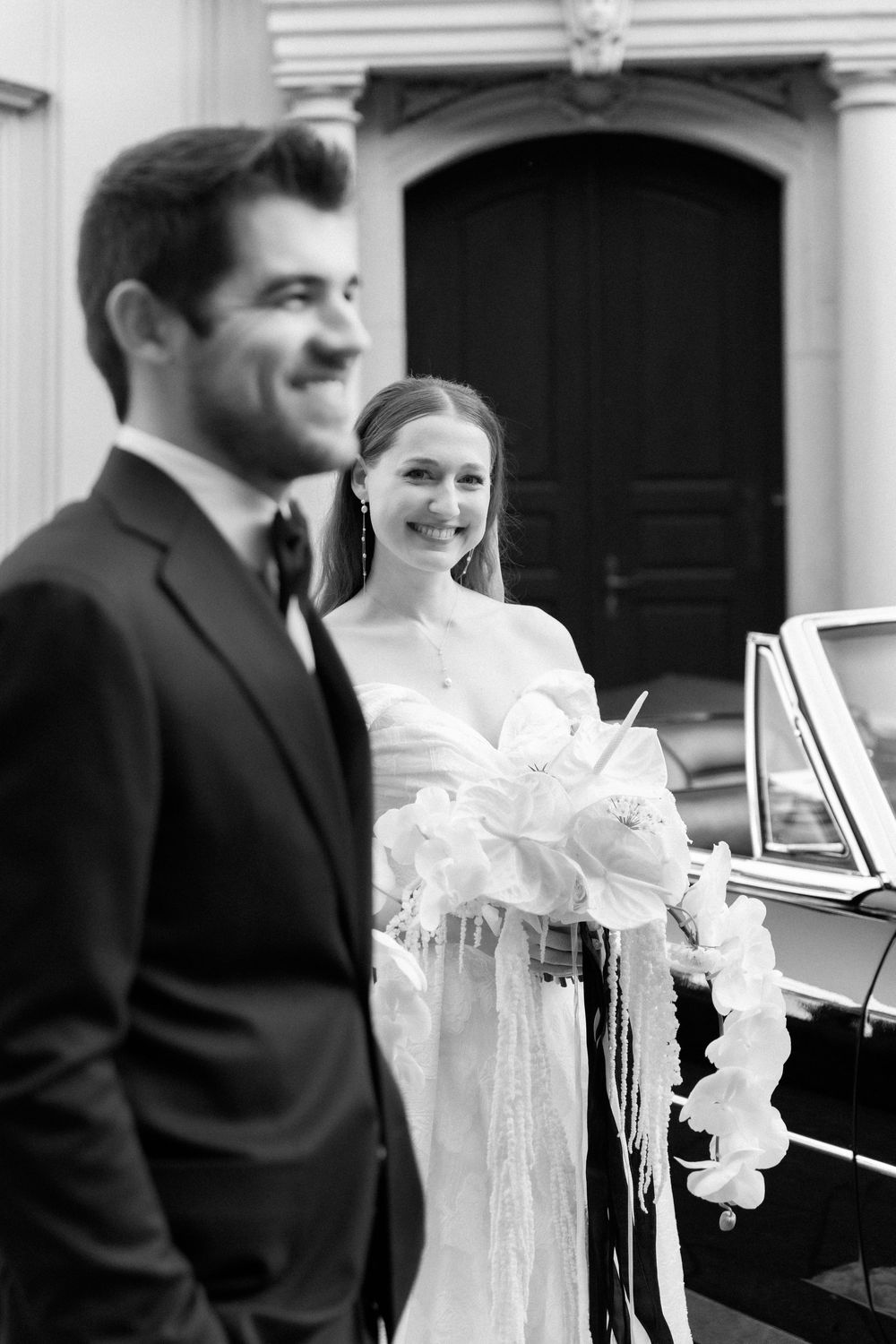 A black and white wedding photo showing a bride in a flowing lace dress standing by a classic car with someone in a suit nearby.