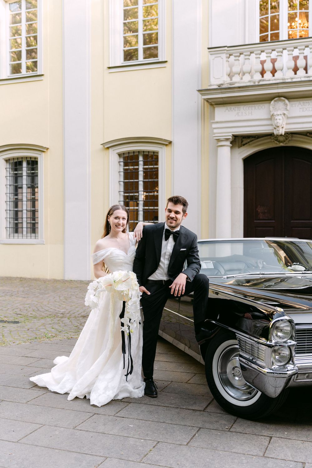 Elegant wedding couple poses beside vintage black Cadillac outside historic cream-colored building with ornate windows.