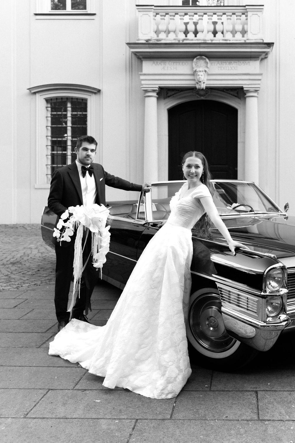 Classic vintage Cadillac car with couple in wedding attire posing outside elegant building in black and white photo.