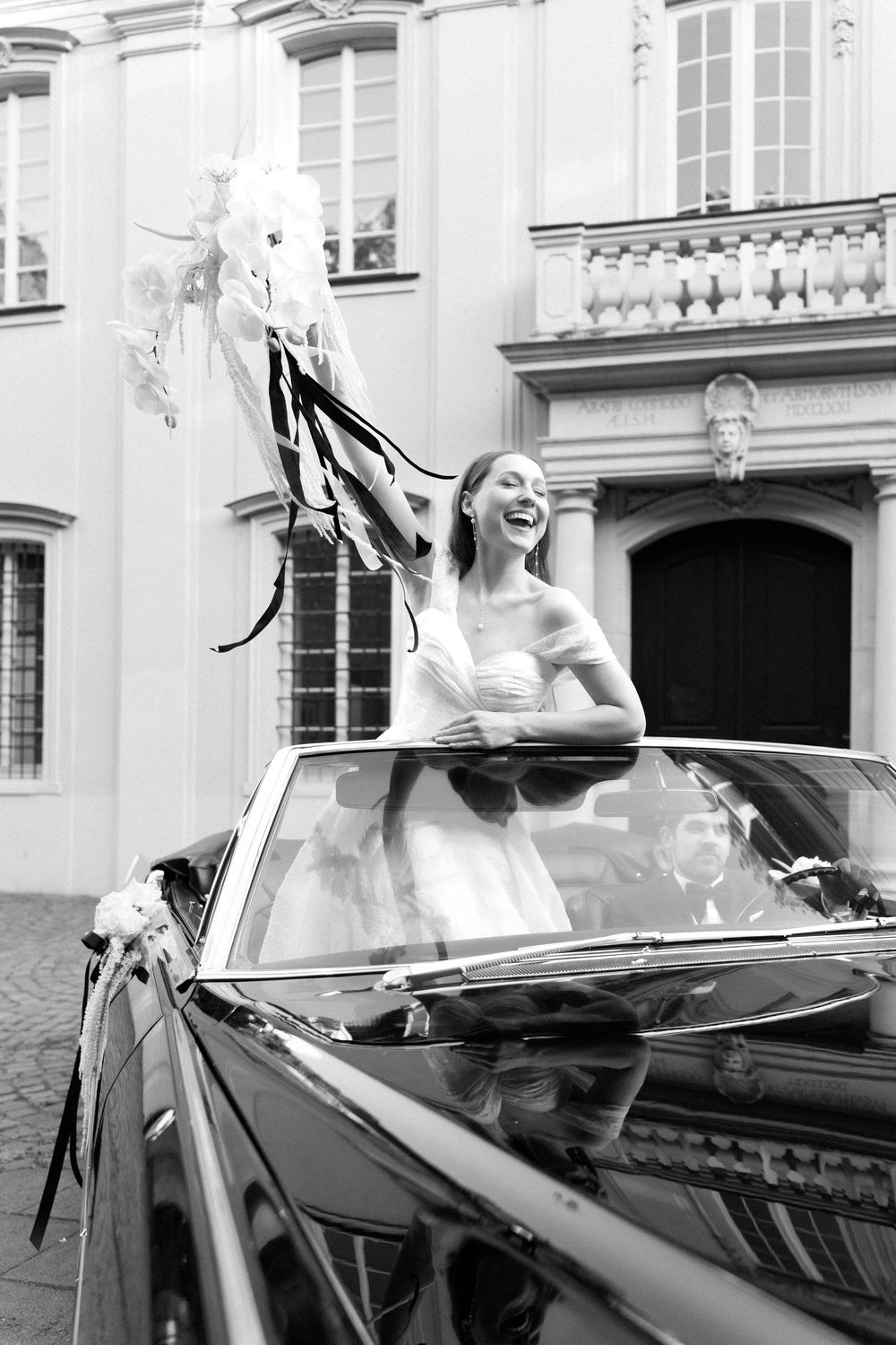 Joyful woman in wedding dress standing up through sunroof of vintage convertible car near elegant building.