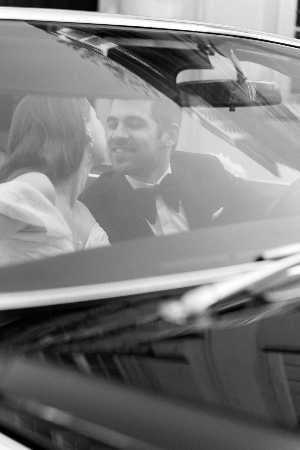 A black and white wedding photo showing a couple sitting together in a car, viewed through the windshield.